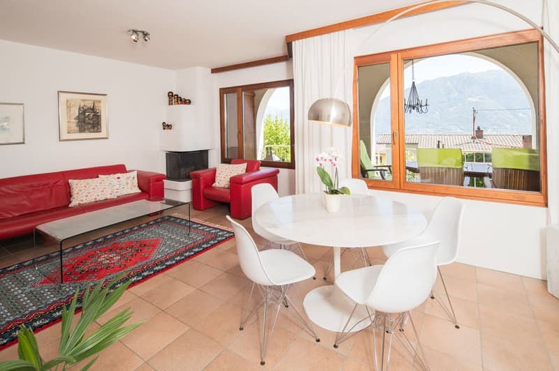 The image shows a spacious living room and dining area with large windows offering a scenic view. The living room features a red sofa and armchairs, a glass coffee table, and a decorative rug. The dining area has a round white table with white chairs. The