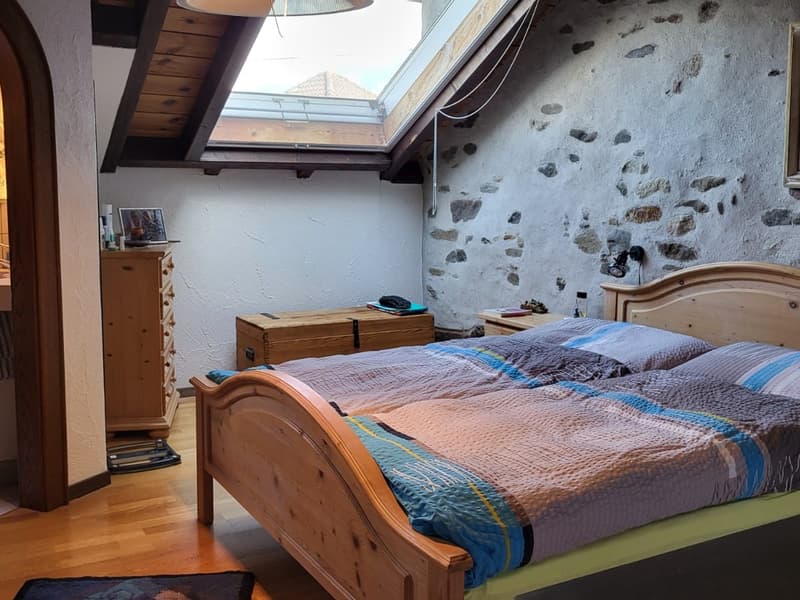 Rustic bedroom with exposed stone wall, wooden beams, and a large skylight. The room features a wooden bed frame, a wooden chest, and a cozy quilt on the bed.
