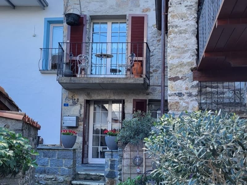 2-story stone building with blue window frames, red shutters, and a balcony on the upper level. The building has a rustic, traditional appearance with potted plants and greenery around the entrance.