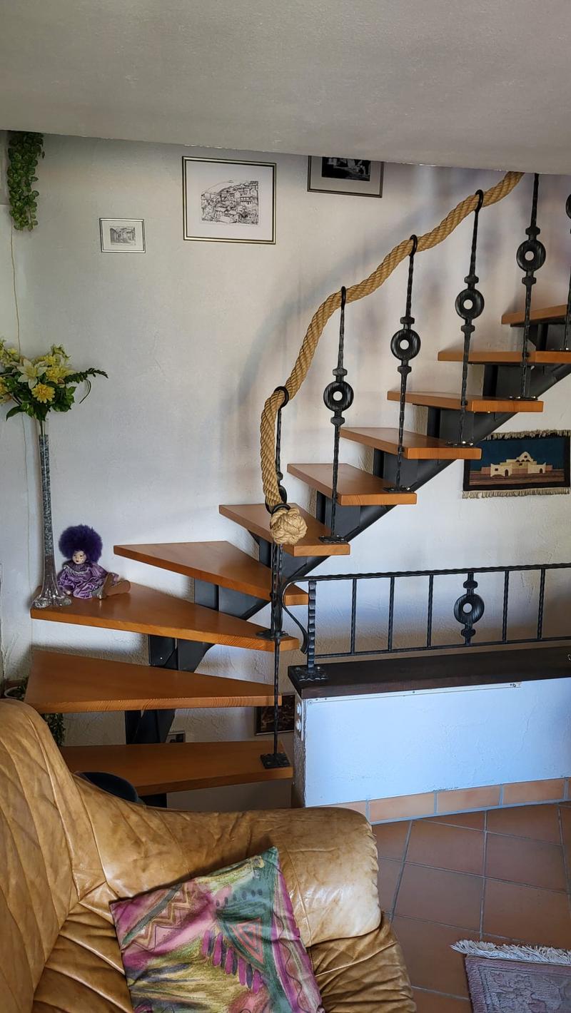 The image shows a living room with a spiral staircase featuring wooden steps and a metal railing with decorative elements. The staircase is accented by thick rope handrails. The room also has a fireplace, a couch with colorful patterned pillows, and some
