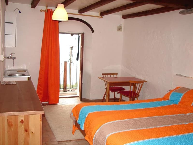 white walls, wooden floor, bed, desk with sink, table and chairs, bedspread with orange, blue, and beige stripes, curtain, lamp, door to balcony