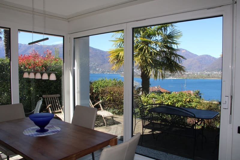 Large windows with a scenic view of a lake surrounded by mountains and palm trees, wooden dining table with chairs, outdoor seating area on the balcony/terrace