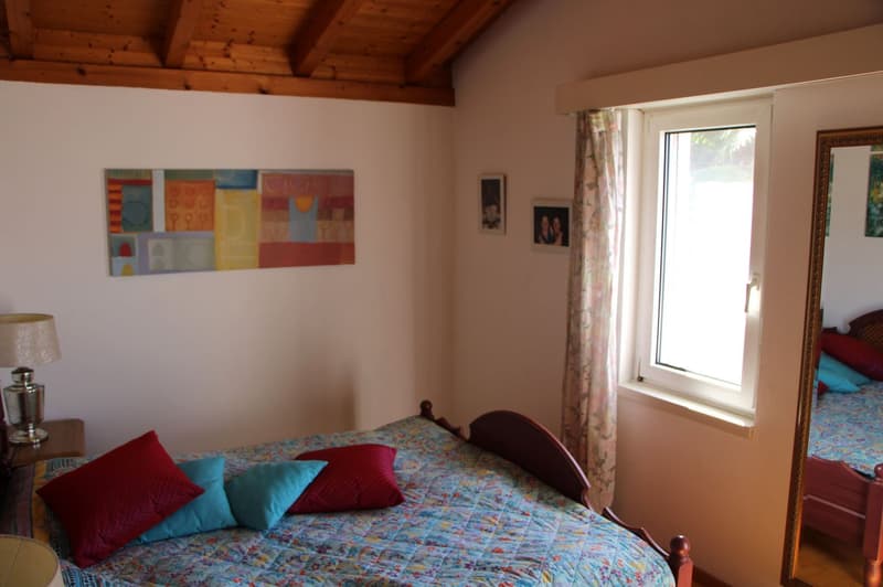 Cozy bedroom with wooden ceiling beams, colorful artwork on the wall, large window, bed with patterned bedding and decorative pillows, side table with lamp
