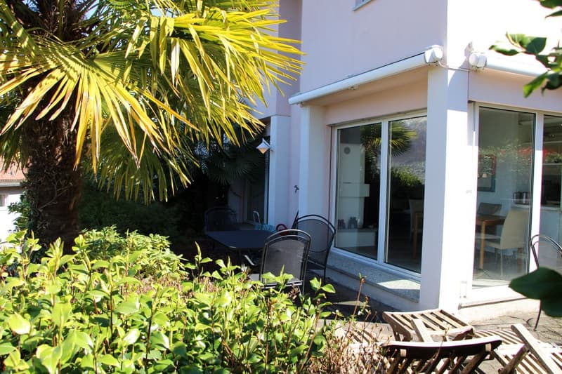 Outdoor patio area with a table and chairs, surrounded by lush greenery including palm trees and other plants. The house has large windows and a sliding glass door leading to the patio.