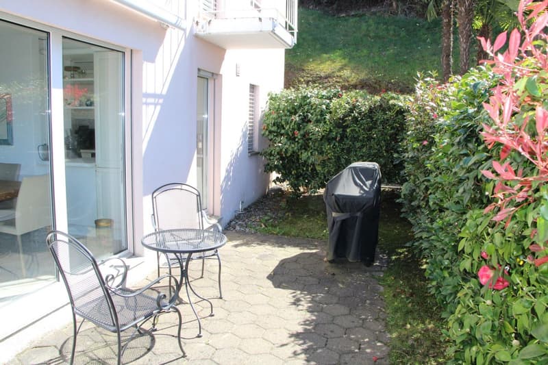 Ground floor apartment with a small patio area, surrounded by lush greenery and vegetation. The patio has a metal table and chairs, and a covered grill or barbecue area. There is also a parked vehicle visible in the image.