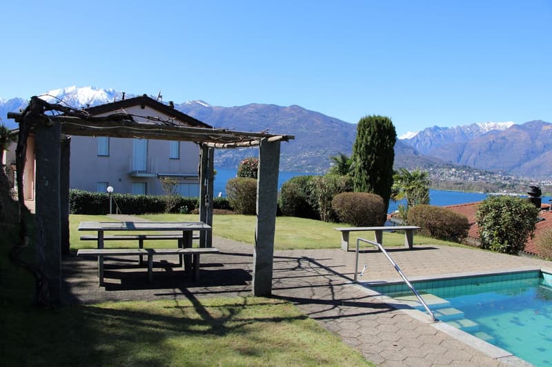 Outdoor patio with picnic tables, swimming pool, surrounded by lush greenery and with a view of the mountains and lake in the background