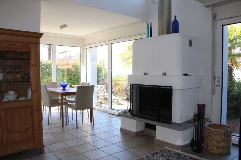 Spacious living room with large windows overlooking a garden, a fireplace, a wooden cabinet, a dining table with chairs, and decorative glass bottles on display.