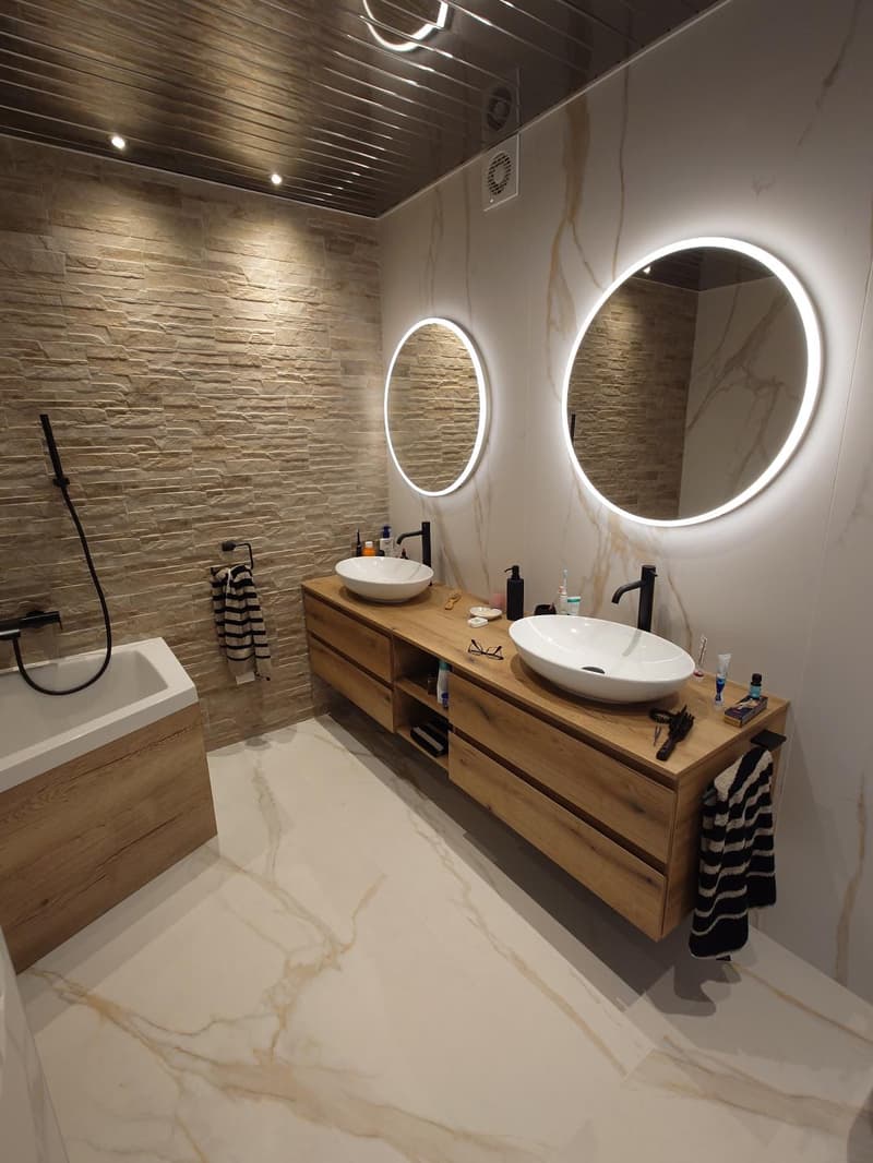 marble tiled floor, white bathtub, large double vanity, two large round mirrors, black faucets