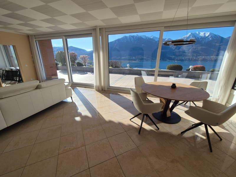modern living room with large windows, beige tiles, white sofa, round wooden table, white chairs, view of mountains and lake
