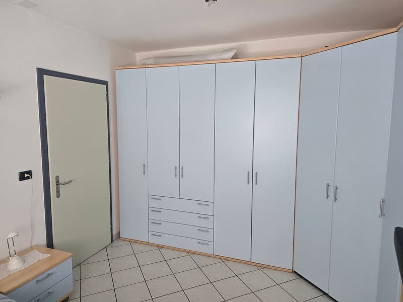 A room with a white wall, a white door, a white cabinet with drawers, a wooden table with a lamp, and a tiled floor.