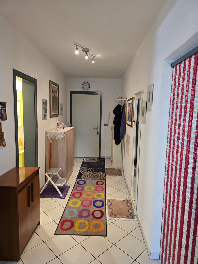 white tiled floor, wooden cabinet, colorful rug, framed pictures on the wall, clock, door, coat rack