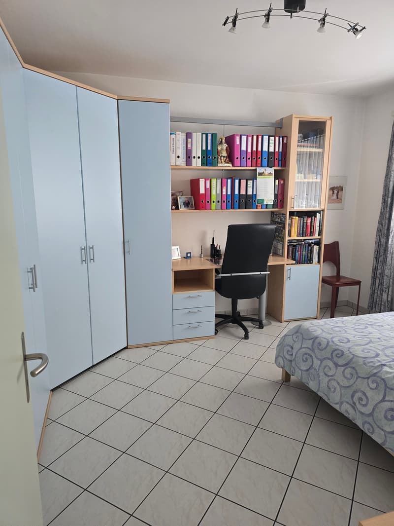 A room with a desk, chair, bed, cabinets, and shelves with books and folders