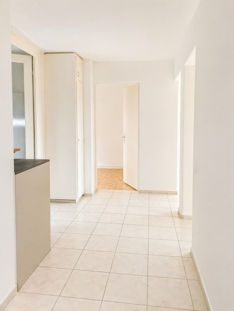 Empty room with white tiles, white walls, wooden floor in the hallway, white cabinet, open door