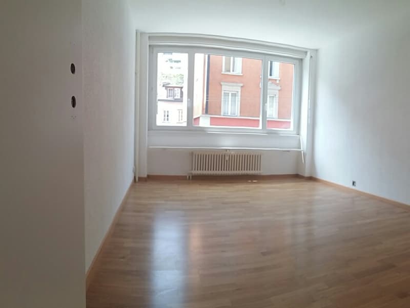 Empty room with wooden floor, white walls, large window, radiator
