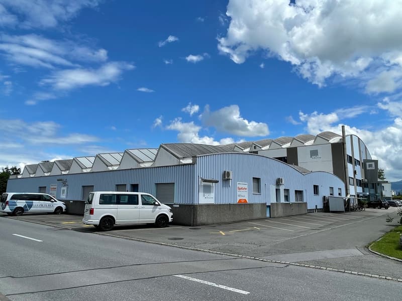 Large industrial or commercial building with a gray metal exterior, multiple entrances, and a parking lot in the foreground. The building has a curved roof design with multiple sections. There are several vehicles parked in the lot.