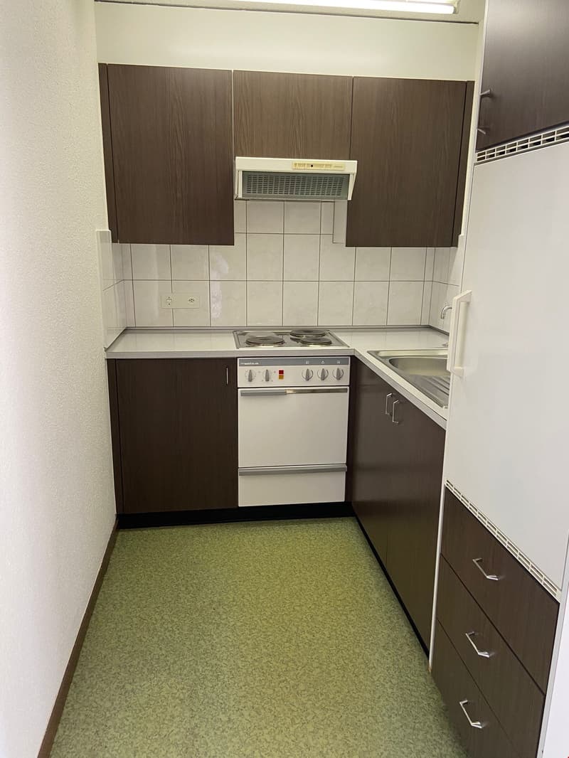 Compact kitchen with dark brown cabinets, white appliances including a stove and dishwasher, tiled backsplash, and green linoleum flooring.