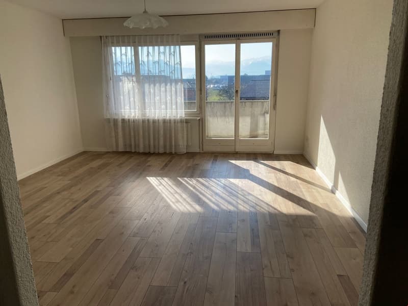 Spacious living room with hardwood flooring, large windows providing natural light, and a sliding glass door leading to a balcony or terrace with a view of the surrounding area.