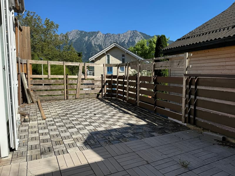 The image shows a wooden deck or patio area with a tiled floor. There are wooden fences and gates surrounding the area, and in the background, there are mountains visible under a clear blue sky. The area appears to be part of a residential property or vac