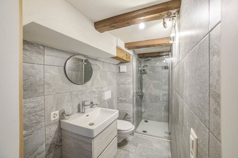 Bathroom with gray tile walls, a white sink vanity, a round mirror, and a glass-enclosed shower stall. The ceiling has exposed wooden beams.