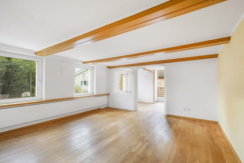 Spacious room with hardwood floors, white walls, and exposed wooden beams on the ceiling. Large windows provide natural light and a view of the surrounding trees.