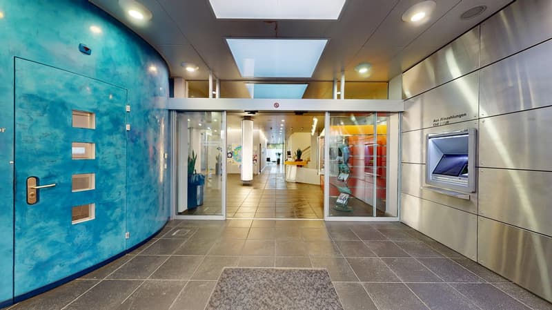 bright lobby, large entryway, double doors, blue wall, automatic sliding doors, carpeted floor, glass walls, white tile flooring, overhead lighting, ATM