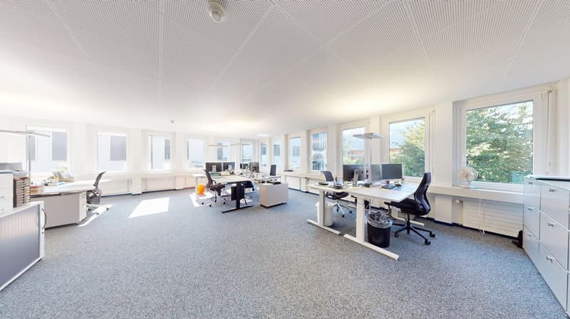 Modern office with neutral color palette. Office desks with computer monitors, office chairs, and trash bins. Large windows with natural light.
