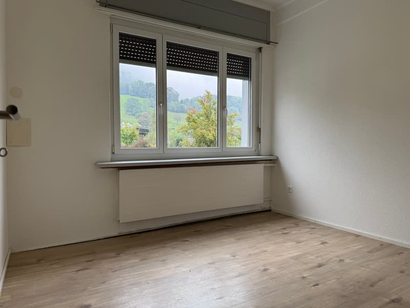An empty room with white walls, wooden floor, a window with a view of the countryside and a window sill with a radiator underneath.