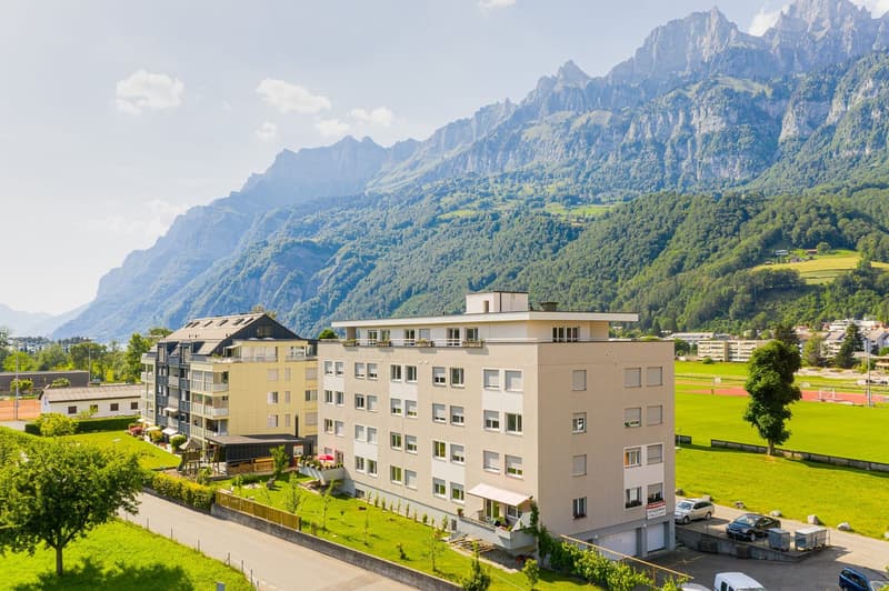 A multi-story apartment building with a parking lot in the foreground, surrounded by lush greenery and mountains in the background. The building has a modern, beige exterior with balconies and windows.