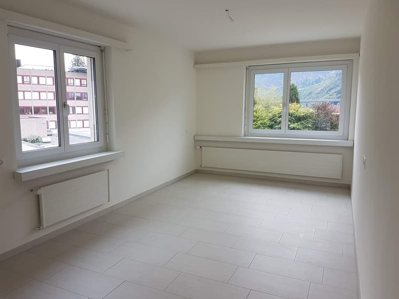 Spacious bedroom with large windows offering a scenic view of the surrounding landscape. The room has white walls and a tiled floor, providing a clean and minimalist aesthetic. There is a radiator installed along the wall, suggesting the room is equipped