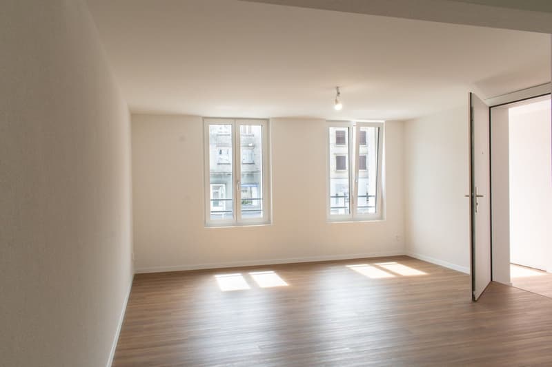 Empty room with wooden floor, white walls, two windows, and an open door.