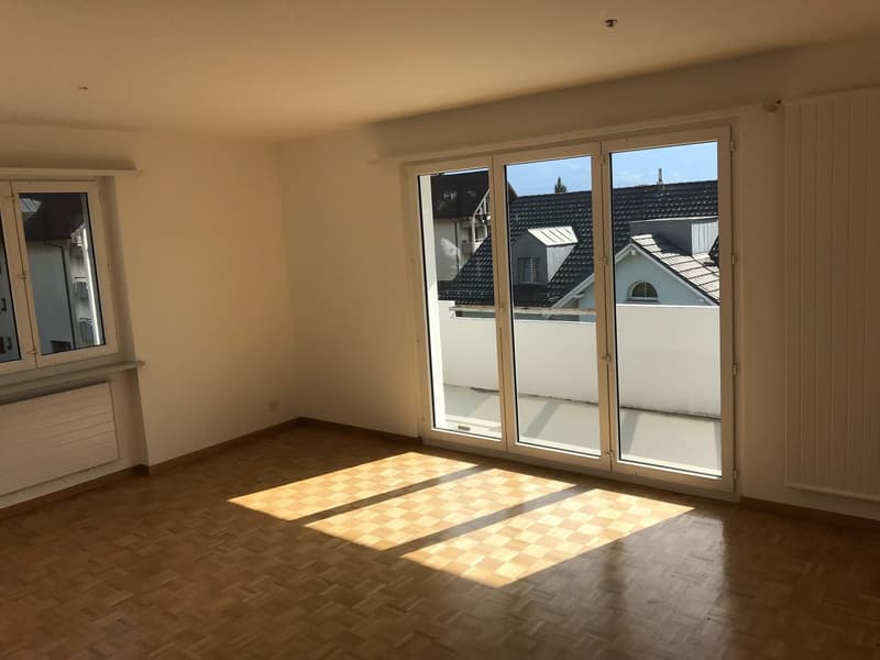 Spacious living room with large windows, hardwood flooring, and a balcony or terrace overlooking the surrounding buildings.