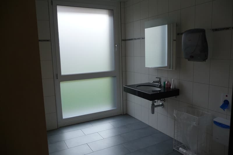 Bathroom with a sink, mirror, trash bin, soap dispenser, hand dryer, and tiled floor.