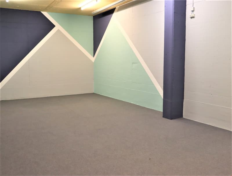 The image shows a large, open room with a concrete floor and painted walls. The walls are painted in a combination of dark blue and light green colors, creating a geometric pattern. There is a support column visible in the room, and the overall space appe