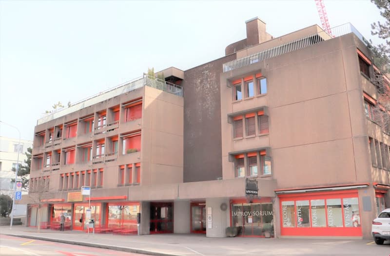 A multi-story commercial building with a modern, angular design. The building has a mix of red and gray tones, with large windows and balconies on the upper floors. There are several retail stores or businesses located on the ground floor.
