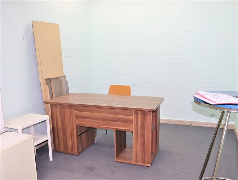 The image shows a simple office setup with a wooden desk, an orange chair, and some storage boxes or shelves on the side. The walls are white, and the floor appears to be a gray carpet or tile. There are no other visible features in the image.