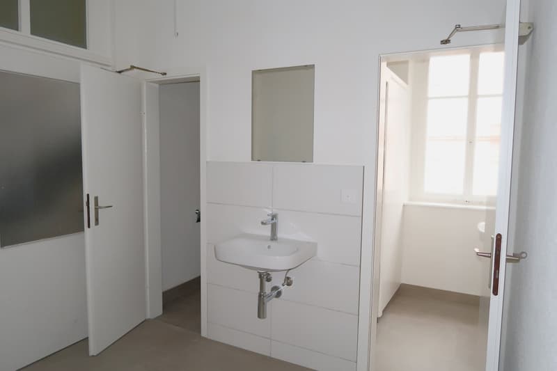 White bathroom with a sink, mirror, and door leading to another room