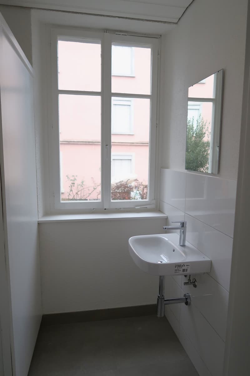 Small bathroom with white walls, a single sink vanity, and a large window overlooking the exterior of the building.