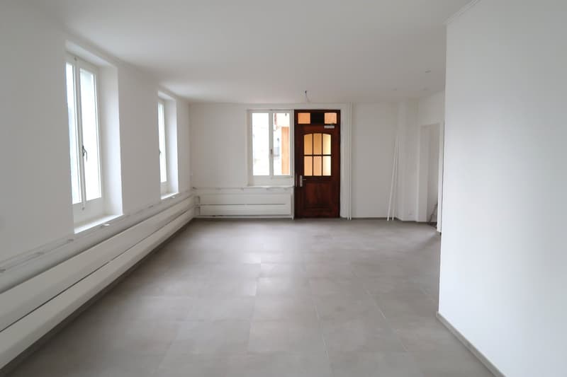 Large open space with white walls, hardwood floors, and a wooden door. The room has large windows providing ample natural light.