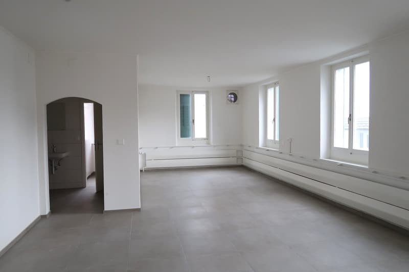 Large open-plan living space with white walls, gray tile flooring, and large windows providing natural light. The room appears to be empty, with no furniture or decor visible.