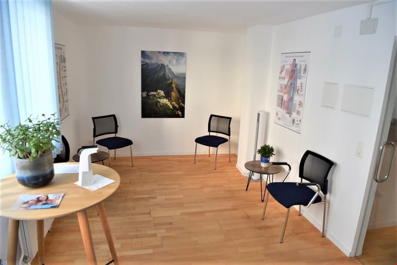 The image shows a small office or consultation room with a wooden floor, white walls, and several pieces of furniture. There are two black chairs facing each other, a small round table, and a plant in the corner. On the wall, there is a large landscape ph