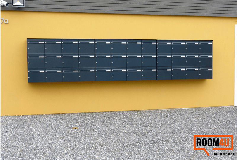 Yellow wall with a row of mailboxes attached to it, gray gravel ground, surveillance camera and light above the mailboxes