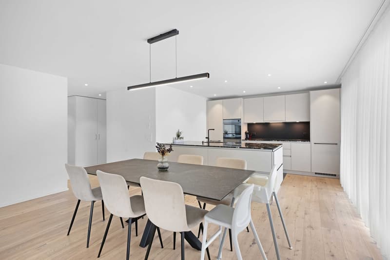 modern dining room with a large dining table, white chairs, white cabinets, wooden floor, pendant light, kitchen island with a sink and oven