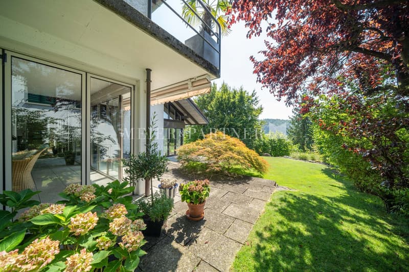 A modern house with glass doors and balcony. It has paved pathway, lush garden with trees and grass. There is a tree trunk forming a natural stone wall behind the house.