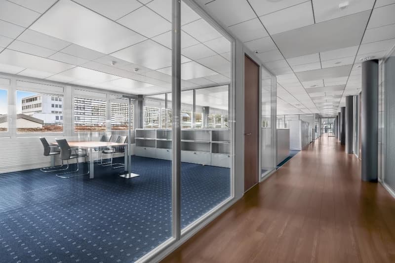 The image shows a modern office space with a conference room visible through glass walls. The office has a long hallway with wooden floors and a blue patterned carpet. The conference room has a large table and chairs, and there appears to be a kitchen or