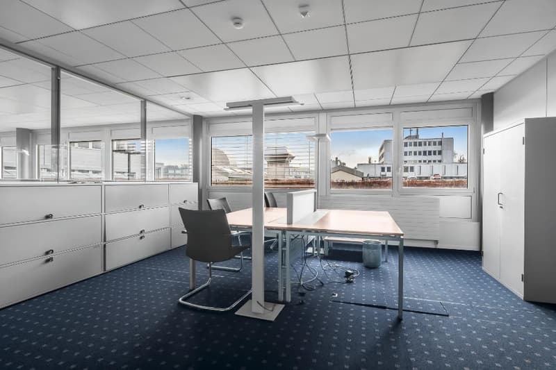 Spacious office with large windows providing a view of the city skyline, modular furniture including desks and chairs, filing cabinets, and a carpeted floor.