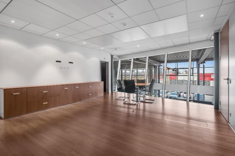 Large open office space with hardwood floors, floor-to-ceiling windows providing a view, built-in cabinets and storage, and a conference table with chairs.