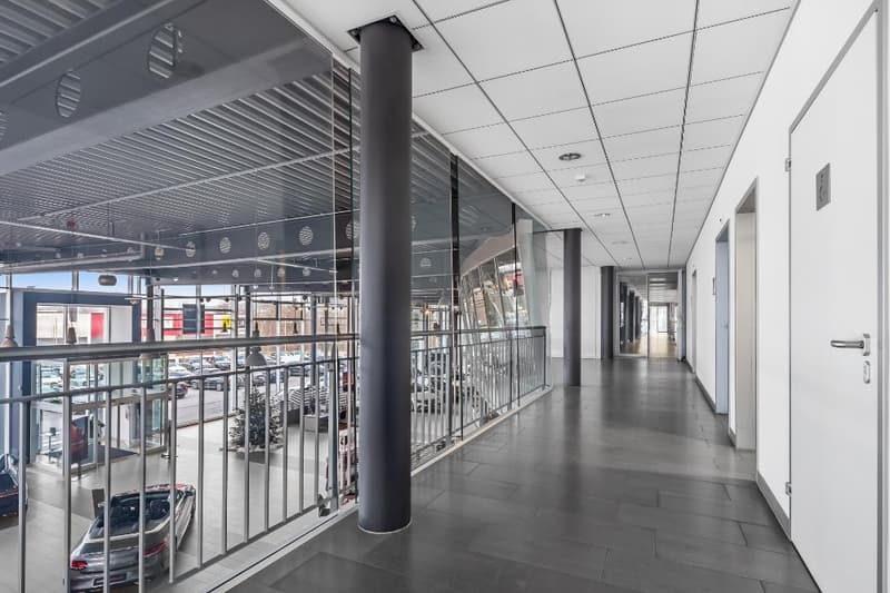 The image shows the interior of a commercial building, with a long hallway or corridor. The hallway has a glass wall on one side, allowing a view of the building's interior spaces. The ceiling is covered with a grid of suspended panels, and the floor is t
