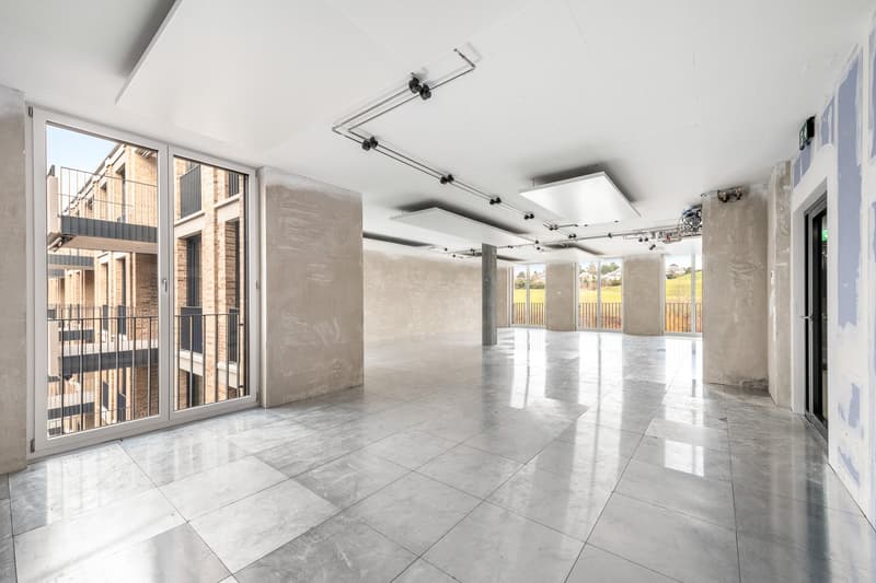 Large open-plan space with high ceilings, concrete walls, and large windows providing natural light. The room has a polished tile floor and track lighting system. There is a balcony or terrace visible through the windows.