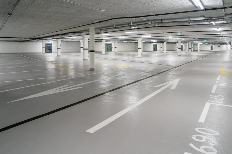 Large indoor parking garage with concrete floors, white walls, and numerous parking spaces. The floor is marked with yellow directional arrows and lines to guide vehicle traffic. The space appears well-lit with overhead lighting fixtures.