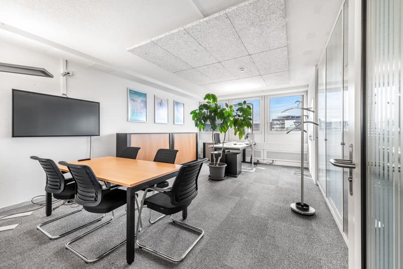 Office room with wooden desk, 6 black office chairs, large mounted television, plant, light grey carpet, picture frames